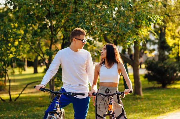 在秋天的公园里 一对年轻貌美的情侣在骑自行车 彼此严肃地望着对方 户外散步时骑自行车的时尚男女的画像 — 图库照片