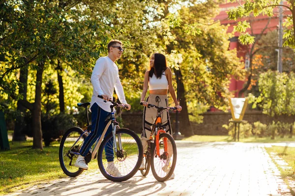 Happy young people in stylish casual clothes riding a bike through a beautiful autumn park. Loving young couple spends active time on bicycles outdoors on a background of sunset.