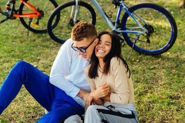 公園の芝生の上に座って楽しみを持っている愛の幸せなカップル 男は女性をくすぐる 彼女は笑顔 自転車を背景に芝生の上でスタイリッシュな若いカップルのレジャー — ストック写真