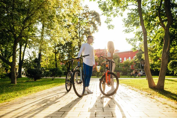 秋の公園で女性と男性とサイクリング後に休憩 夕暮れ時に公園を自転車で歩くスタイリッシュな美しいカップル — ストック写真