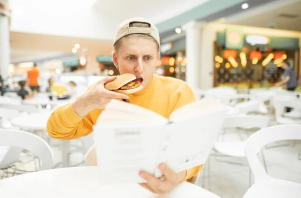 모자와 주황색 스웨터를 청년이 패스트푸드 점에서 햄버거를 표정이 학생이 공부하면서 — 스톡 사진