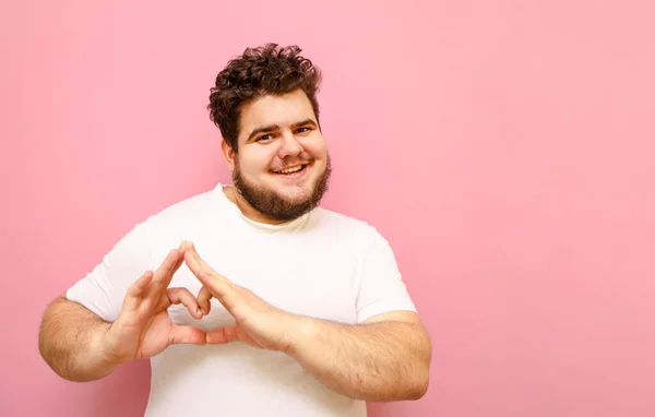 Happy fat man in a white T-shirt and with a beard stands on a pink background, shows a gesture of heart and looks into the camera with a smile on his face. Curly overweight guy showing love gesture