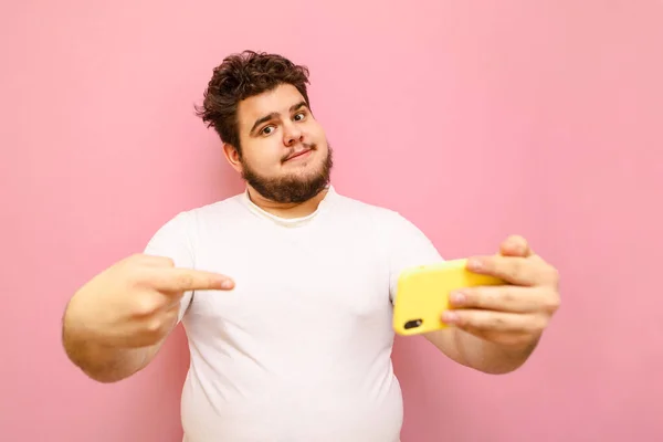 Handsome young man with beard and curly hair looks into the camera and points his finger at the smartphone in his hand. Fat man in white t-shirt stands on pink background with smartphone in hand