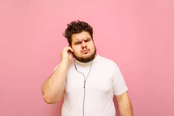 Funny bearded young fat man with displeased face listening to music in headphones and looking into camera. Cheerful guy with overweight and curly hair in headphones isolated on pink background.