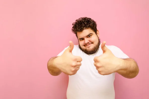 Cheerful bearded fat man in white t-shirt isolated on pink background, looks into camera and shows thumbs up. Positive guy with overweight and curly hair shows gesture like.