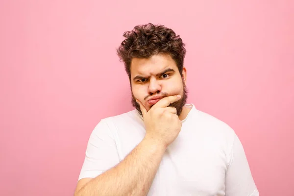 Funny fat man in white t-shirt looks into camera with pensive serious face on pink background, isolated. Funny pensive guy with overweight, beard and curly hair, isolated.