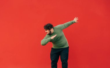 Sıradan giyinmiş, kıvırcık saçlı, kırmızı duvarda dans eden ve gülümseyen pozitif şişman bir adam. Şişman adam kırmızı üzerine izole edilmiş bir meşe ağacı gösteriyor..