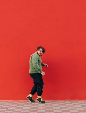 Kırmızı duvarda dans eden ve kameraya bakan şık sokak kıyafetleriyle dans eden şişman adamın dikey fotoğrafı..