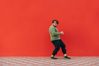 Karizmatik, şişman, yeşil kazaklı genç adam kırmızı arka planda hip hop yapıyor ve kablosuz kulaklıkla müzik dinliyor..