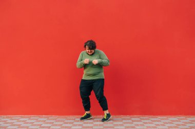 Yeşil kazaklı komik şişman adam kırmızı bir duvarın arkasında sokakta dans ediyor. Kırmızı arka planda dans eden kilolu adam..