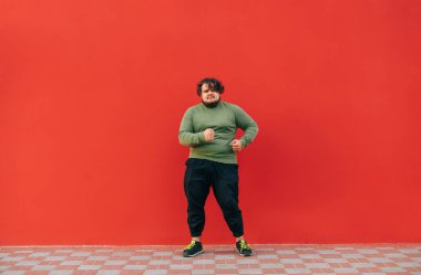 Komik, şişman bir adam kırmızı arka planda dans ediyor ve yüzünde bir gülümsemeyle başka yere bakıyor. Dans eden şişman bir adamın tam boy portresi..