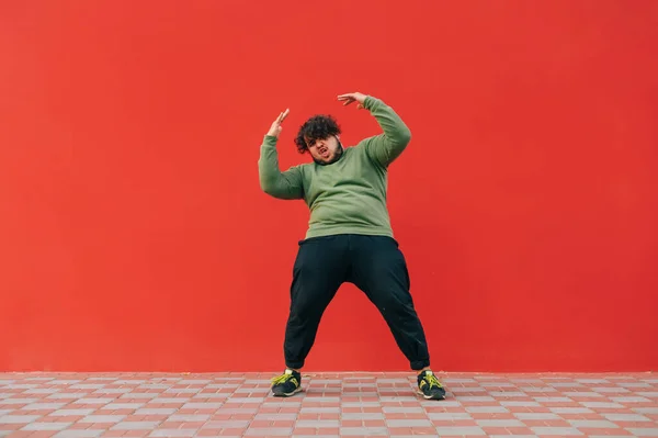 Funny fat guy in casual clothes dances on a red wall and looks at the camera. Overweight man shows a dancing performance.