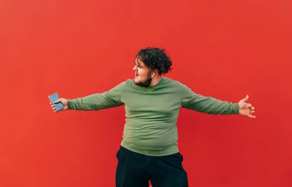 Funny fat man with curly hair dancing on a red background with a smartphone in his hands wireless headphones in his ears. Isolated.