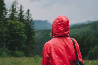 Kırmızı yağmurluk giymiş, sırt çantalı, dağ zincirine ve dağ köknarı ormanının pitoresk manzarasına bakan bir kadın yolcunun arka görüntüsü..