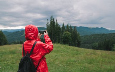 Kırmızı ceketli, akıllı telefonunu kullanan ve dağ tepeleri ve ormanlar gibi güzel bir dağ manzarasının fotoğraflarını çeken bir kadın yolcunun arka planı..