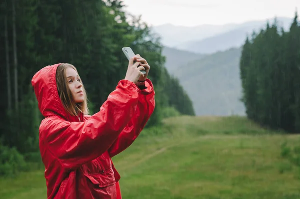 Side view of a hiker woman taking selfie on her mobile phone, wearing a red raincoat in the pouring rain in the mountain forest. Wet weather. Technology, travel concept.