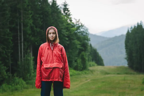 若いかなり観光客の女性は 夏の雨の下で赤い防水ジャケットで山の丘の上の写真のポーズをとっています 雨の多い日 — ストック写真