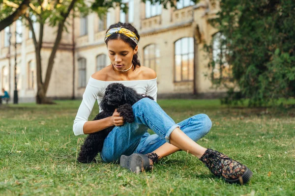 公園の芝生の上に座っている美しいヒスパニック系の女性の肖像画彼女の腕の中に黒い犬と遊んでいる建築の背景に — ストック写真
