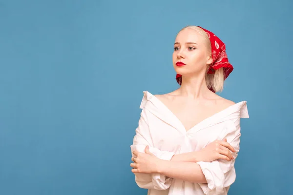 Attractive lady in white men's shirt and headscarf isolated on blue background, looking away with serious face. Girl peasant in shirt and red handkerchief posing on camera, looks at copy space