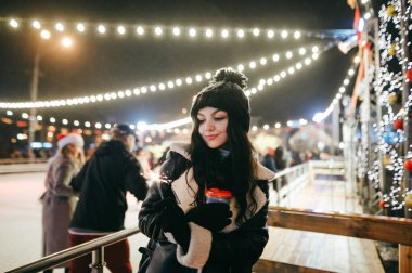 Şirin bayan elinde maytaplar ve arka planda bir fincan sıcak içecekle Noel sahasında duruyor ve gülümsüyor. Kız Noel 'i panayırda geçiriyor..