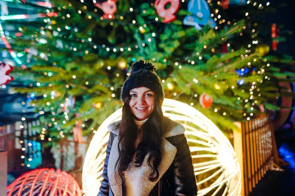 Happy girl in warm clothes stands on the street on the background of a decorated Christmas tree and looks at the camera with a smile on his face. Walk at Christmas. New year concept.