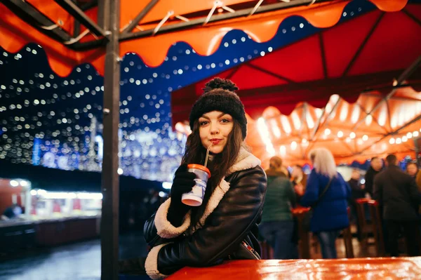 暖かい服のかわいい女の子はクリスマスマーケットで夕方に暖かい飲み物を飲み カメラを見ています 女性は冬のフェアで時間を過ごす — ストック写真