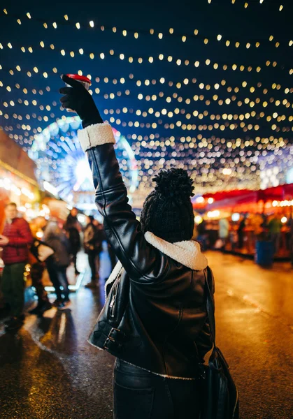 冬服の女性は 彼女の手に暖かい飲み物のカップとクリスマスマーケットで夜に立っています 提灯で飾られた通りの背景からの眺め — ストック写真