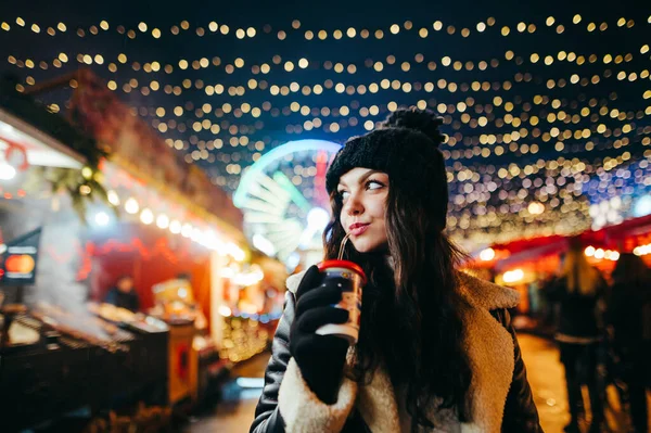 帽子のかわいい女性と暖かい服の肖像画は 美しい装飾が施されたクリスマスの夜のフェアの背景に暖かい飲み物を飲んでいます 冬のお祝いの街を歩く女の子 — ストック写真