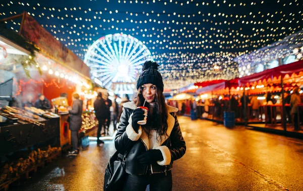 暖かい服を着た素敵な女性の肖像画は 美しい光の装飾が施されたクリスマスマーケットに立って 熱い飲み物を飲み 離れて見ています 街のクリスマスセンターの周りを夜歩く — ストック写真