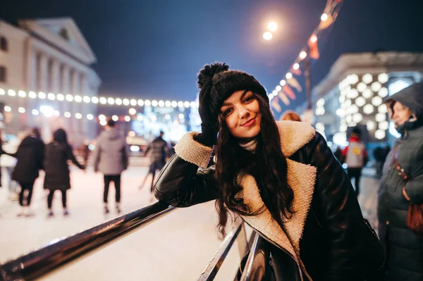 かわいい女性は アイスリンクやポーズ クリスマスの装飾された通りの背景の近くの通りの夜に立って カメラと笑顔に見えます クリスマス街の少女の夜の肖像画 — ストック写真