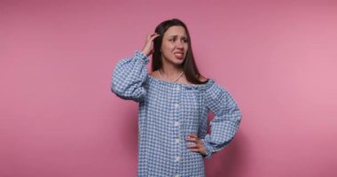 Bir kadın pembe bir arka fonda duruyor mavi bir gingham elbise giyiyor. Elleriyle soru sorarak hareket ediyor, karışık duygular sergiliyor, neşe içinde poz veriyor..