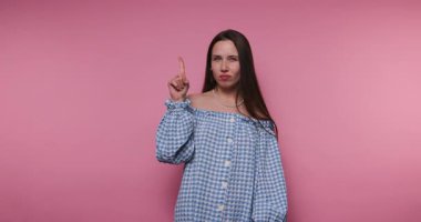 Bir kadın pembe bir zemine karşı duruyor ve ciddi bir jest yaparak parmağını kaldırıyor. Mavi beyaz kareli bir bluz giyer, güçlü duygular gösterir..
