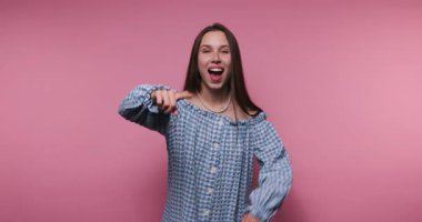 Genç bir kadın canlı pembe bir fon karşısında heyecanla gülümser ve el kol hareketi yapar. Neşeli enerjisi ve şık kıyafeti eğlenceli, neşeli ve bulaşıcı bir atmosfer yaratıyor..