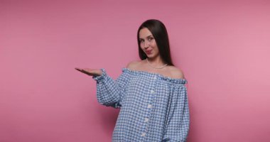 Genç bir kadın canlı pembe bir arkaplan önünde dikilirken şık damalı kıyafetini sergiliyor. Rahatlamış bir tavır sergiliyor. Kendine güvenen bir gülümsemesi ve neşeli bir el hareketi var..