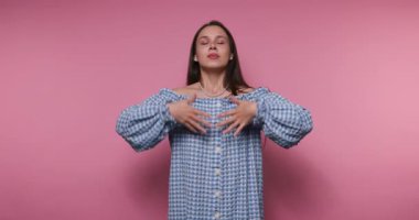 Bir kadın, gözleri kapalı ve canlı pembe bir fon karşısında nazikçe ellerini kaldırarak güvenle duruyor. Şık, mavi, kareli bir elbise giyiyor, sükunet ve soğukkanlılığı simgeliyor..