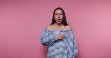 Bir kadın pembe bir duvarın önünde, şaşırmış bir ifadeyle duruyor. Omzunun dışında bir şey giyiyor ve jestler yapıyor. Muhtemelen çevresindeki beklenmedik bir şeye tepki veriyor..