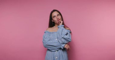 Mavi gingham elbise giyen bir kadın canlı pembe bir arkaplan karşısında duruyor, neşeli bir ifadeyle düşünüyor. Yaratıcı ve eğlenceli bir atmosferi var..