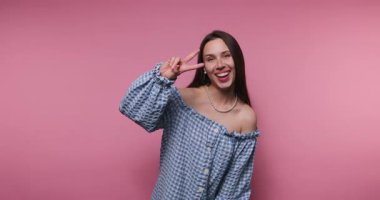 Zarif bir damalı kıyafet giyen genç bir kadın barış işareti ile poz verirken ışıl ışıl gülümsüyor. Canlı pembe arka plan onun neşeli tavrını ve neşeli mizacını arttırır..