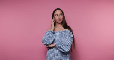 Omuzları kapalı bir şekilde giyinmiş genç bir kadın akıllı telefonuyla neşeli bir şekilde konuşuyor, canlı pembe bir arka plana karşı heyecan ve merak gösteriyor..