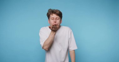 Genç bir adam, mavi arka planda yüzünde bir gülümsemeyle kameraya öpücük yolluyor..