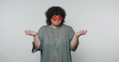 Kıvırcık saçlı bir kadın parlak turuncu güneş gözlüğü takarken neşeli bir ifade sergiler. Minimalist bir stüdyo ortamında duruyor, bir kafa karışıklığı ya da kararsızlık hissi veriyor..