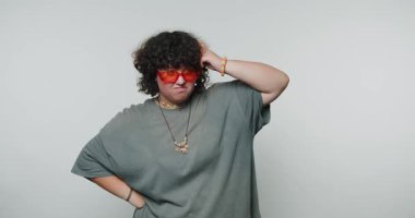 Kıvırcık saçlı ve parlak turuncu gözlüklü bir insan basit bir arkaplanda dururken karışık bir yüz ifadesi sergiler. Günlük kıyafetleri ve eşsiz tarzları neşeli bir havayı vurguluyor..