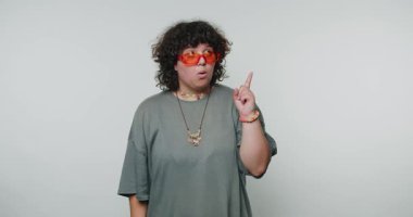 Kıvırcık saçlı ve turuncu güneş gözlüklü bir insan parlak bir stüdyoda fikirleri paylaşırken heyecan verici hareketler yapar. Kıyafetleri gündelik, modaya uygun bir tarzı yansıtıyor..