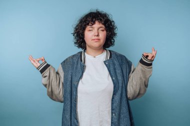 Kıvırcık saçlı genç bir kadının portresi, mavi arka planda kot ceketle huzurlu görünürken meditasyon yapıyor.