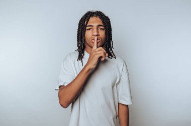 Örgülü saçlı bir gencin portresi, beyaz arka planda sükunet ve gizliliği vurgulayarak sessizlik için bir jest yapar.