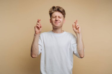 Genç adamın portresi, bej arka planda umut verici bir ifade sergileyerek parmaklarını çapraz yapar.