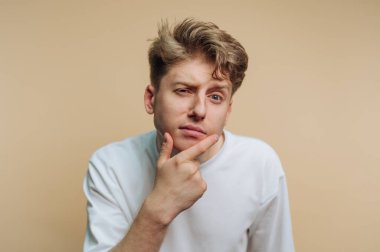 Genç adamın portresi, çenesine dokunurken düşünceli bir görünüm sergiliyor. Bej arka planda merak ve iç gözlem içeriyor.