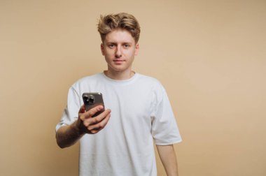 Genç bir adamın portresi, elinde akıllı bir telefonla, bej arka planda sıradan bir stil ve neşeli bir ifade sergiliyor.