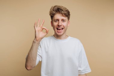 Gülümseyen ve el hareketini gösteren genç bir adamın portresi, pozitifliği somutlaştıran ve bej arka planda onay duygusu