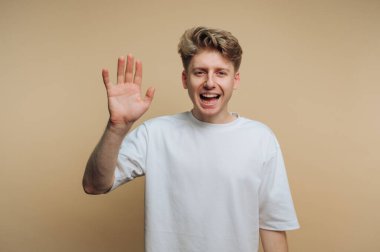 Genç ve neşeli bir adamın portresi enerjik bir şekilde elini sallıyor, canlı bir kişilik ve bej arka planda sıcak bir görünüm sergiliyor.